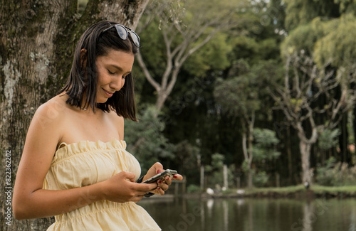 joven mujer hispanoamericana sonríe al escribe mensaje de texto en el teléfono celular al aire libre en el parque  photo