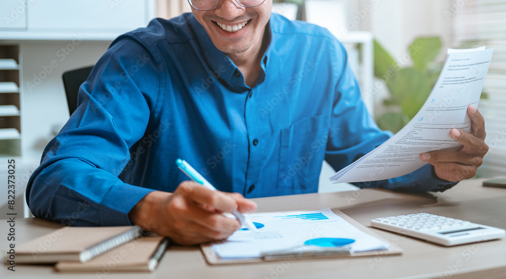 accounting, analyzing, analysis, cost, payment, management, calculate, chart, document, graph. A man is sitting at a desk with a blue shirt on and a pen in his hand. He is smiling and he is happy. accounting, analyzing, analysis, cost, payment, management, calculate, chart, document, graph. A man is sitting at a desk with a blue shirt on and a pen in his hand. He is smiling and he is happy.