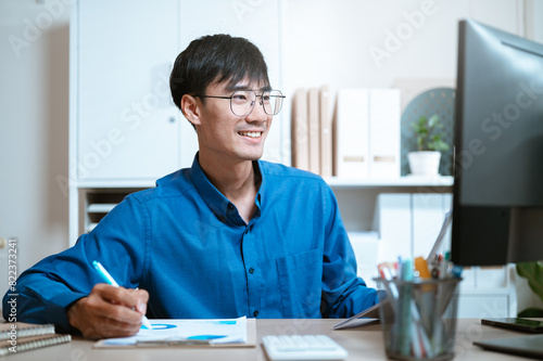 document, thinking, working, writing, executive, expertise, ideas, planning, job, management. A man is sitting with a blue shirt on and writing with a blue pen. He is smiling and enjoying himself.