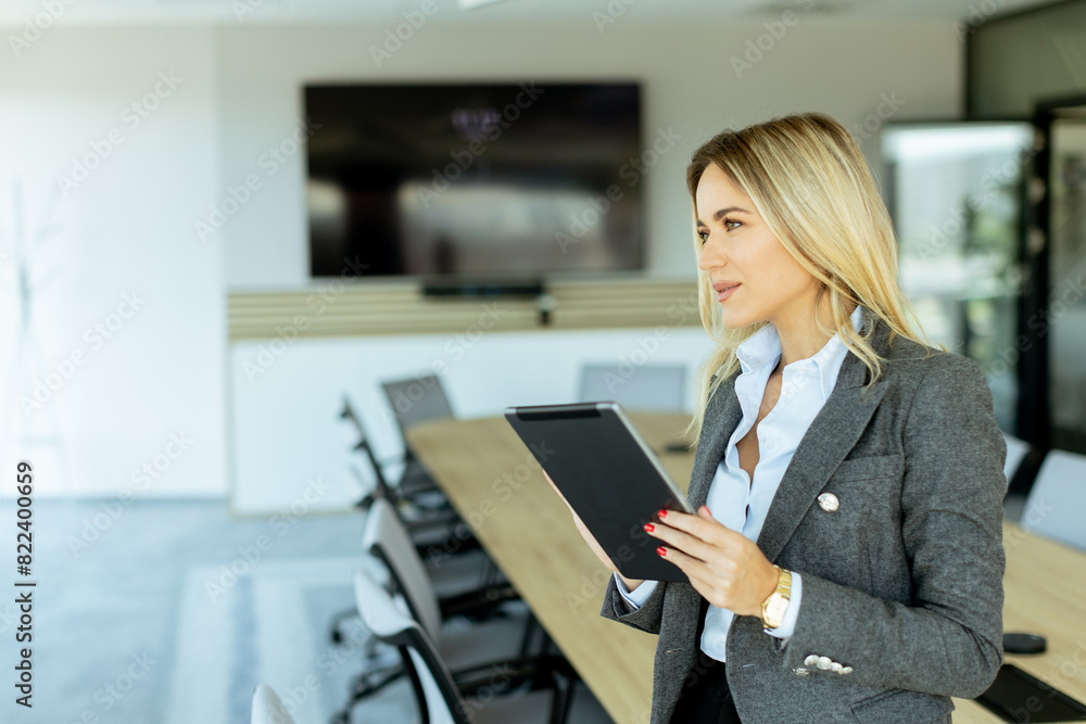 Obraz premium Smiling professional woman in red blazer holding tablet in modern conference room during daytime
