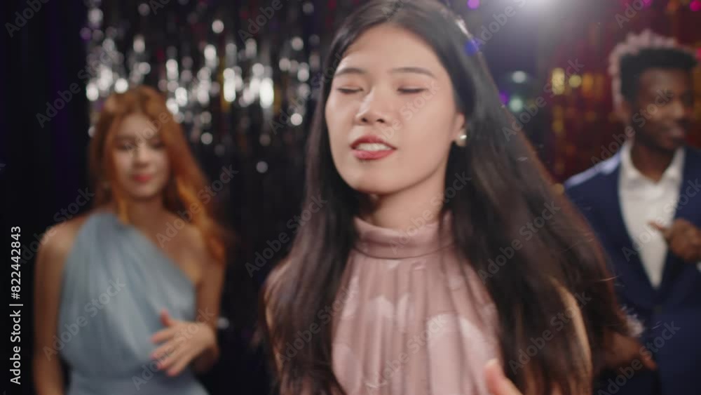 Ecstatic Chinese girl in pink evening gown and white pearl earrings dancing and singing at prom party in front of happy intercultural friends enjoying clubbing together Ecstatic Chinese girl in pink evening gown and white pearl earrings dancing and singing at prom party in front of happy intercultural friends enjoying clubbing together