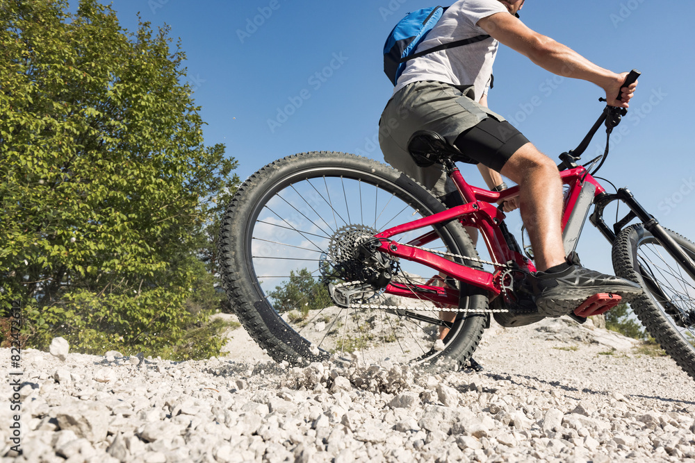 Obraz premium Mountain bike rider riding an electric bike along a white gravel trail on a sunny summer day. Outdoor sports activity concept.