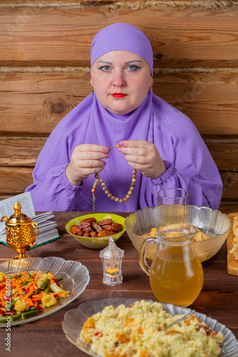 A Muslim woman in a lilac hijab is fingering her rosary at the table, praying for the end of Ramadan