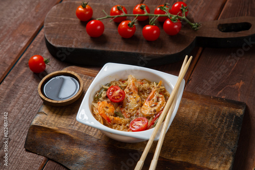 Rice with shrimp in Thai style sos pad thai on a wooden stand next to cherry tomatoes, soy sauce and chopsticks