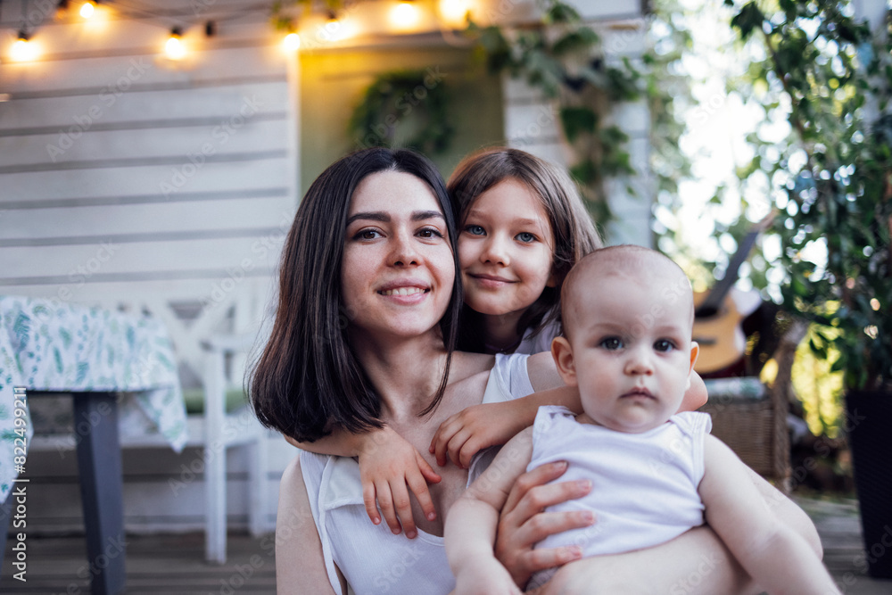 Beautiful smiling brunette mom with two cute little kids. Happy family is hugging on vintage style terrace of wooden house. Beautiful smiling brunette mom with two cute little kids. Happy family is hugging on vintage style terrace of wooden house.