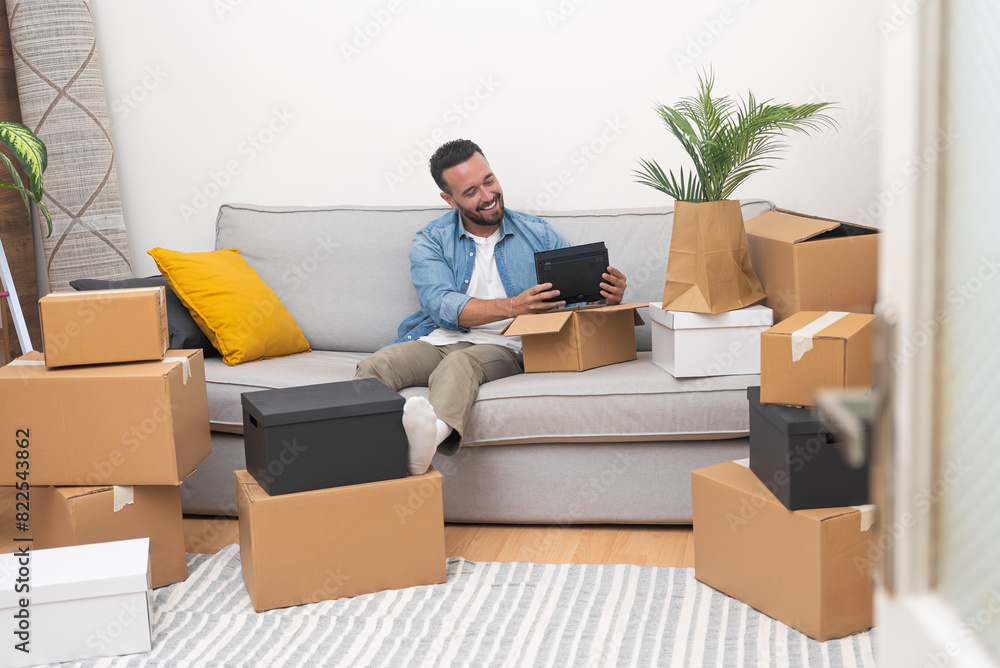 Happy man with delighted expression sits on cozy sofa placing boxes with accessories for home improvement next to each other communicating online with friend Happy man with delighted expression sits on cozy sofa placing boxes with accessories for home improvement next to each other communicating online with friend