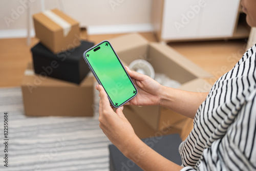 Seamless Move-In: A woman holds a smartphone with a green screen, set against a blur of cardboard boxes in her modern spacious flat during a home relocation