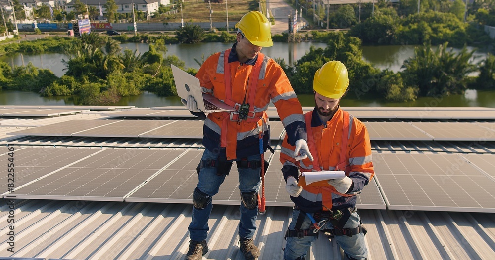 Two engineers solar technicians in reflective gear discussing working examine solar installations on rooftop panels at dusk, with suburban backdrop Two engineers solar technicians in reflective gear discussing working examine solar installations on rooftop panels at dusk, with suburban backdrop