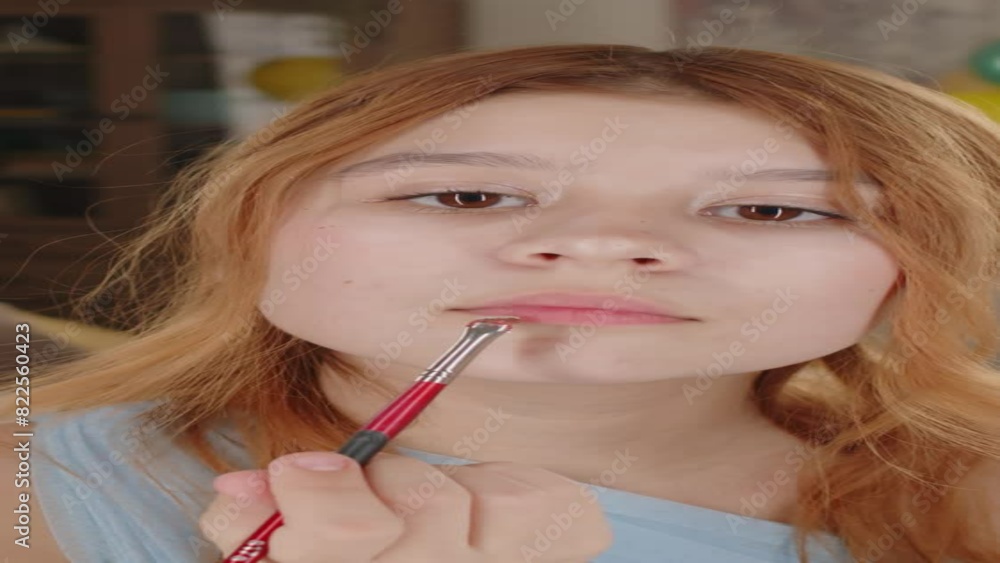 Vertical shot of pretty teenage girl standing in front of mirror and applying makeup on her eyes, eyebrows and lips with cosmetic brush while preparing for prom party Vertical shot of pretty teenage girl standing in front of mirror and applying makeup on her eyes, eyebrows and lips with cosmetic brush while preparing for prom party