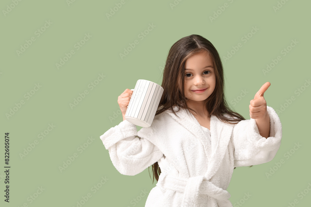 Cute little girl in bathrobe with cup of tea showing thumb-up gesture on green background Cute little girl in bathrobe with cup of tea showing thumb-up gesture on green background