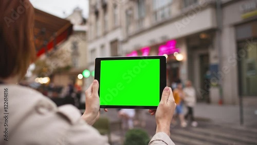 View from behind of Caucasian woman with red hair holding small tablet with both hands. Chroman key on screen of device. Green screen. Girl holding digital device steadily without moving. photo