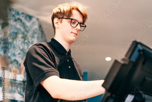 Young waiter serving customer at cash point in cafe. Man working with POS terminal. Cashier, barista checking for payment receipt. Hospitality, server and preparing a slip at the till in coffee shop