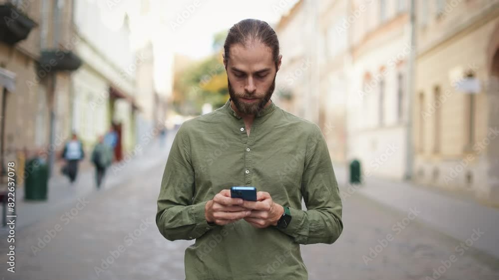 Close frontal focus on Caucasian male with beard standing in middle of street. Using mobile device with both hands. Typing with thumbs on screen of smartphone. Sunny weather. Texting. Close frontal focus on Caucasian male with beard standing in middle of street. Using mobile device with both hands. Typing with thumbs on screen of smartphone. Sunny weather. Texting.