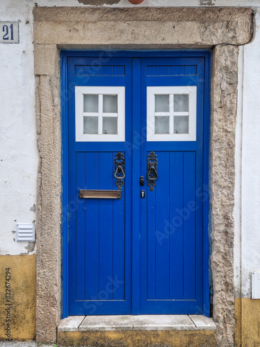Traditional wooden door painted in vibrant blue with vintage knockers and two small hatches on top. Facade of Portuguese typical house