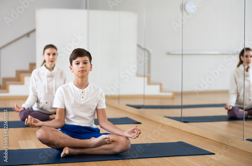 Concentrated teen boy sitting on mat in fitness center, making yoga meditation in lotus pose while exercising with family..