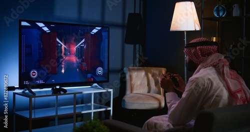 Arabic man playing singleplayer videogames on TV, relaxing in living room. Middle Eastern gamer enjoying science fiction shooter game on gaming console, having fun, handheld camera shot photo