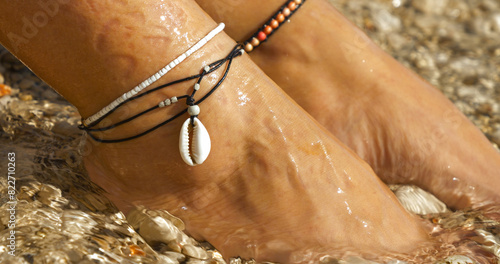 Fotografie CLOSE UP: Elegant anklets with a seashell on female feet by the sparkling sea, reflecting a perfect blend of style and summertime serenity on a pebble seashore