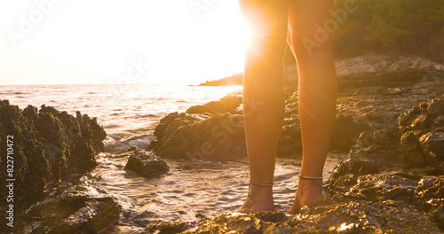 LENS FLARE, CLOSE UP: Lady admires golden sunset barefoot on a seashore where warm rays of setting sun reflect off the wet rocks. She is wearing simple and beautiful shell bracelets around her ankles. photo