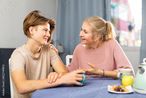 Happy family of mother and teenage son enjoying conversation, sitting at kitchen at home