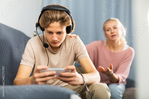 Teenage boy absorbed in online game on mobile phone, ignoring frustrated mother talking to him. Concept of phubbing and internet gaming addiction photo