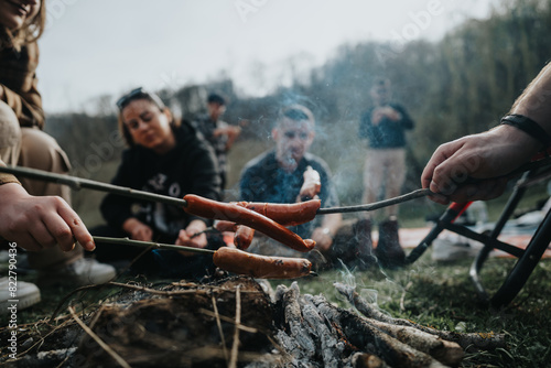 Group of friends gathered around a campfire, roasting hotdogs on sticks, enjoying an outdoor cooking activity together.