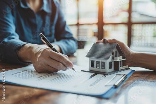 A real estate agent is pointing at a house model on top of a contract while their customer holds a pen to sign the document.