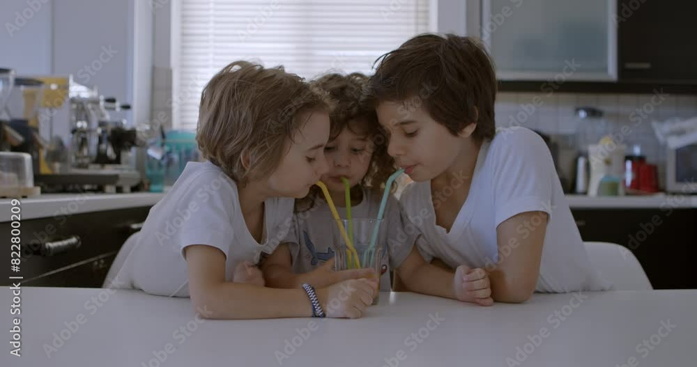 Three children gather around a kitchen table sharing a glass of orange juice with straws in a modern kitchen. Three children gather around a kitchen table sharing a glass of orange juice with straws in a modern kitchen.