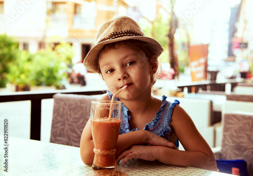Cute calm thinking kid girl in summer hat drinking tasty juice in street restaurant. Closeup