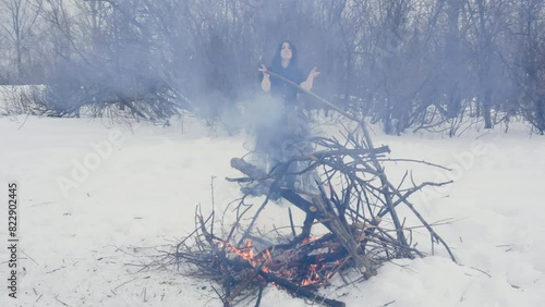 Woman witch near burning bonfire in snowy woodland perform ritual dances in black dress. Ancient mystical spiritual dancing ceremony in winter forest. Scary wizard making ordinance standing on snow.