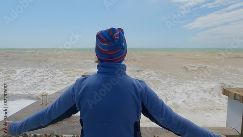 Woman in hat admiring sea storm with huge waves on sandy beach our of season rising hands up. Travel tourism vacations. Female enjoying nature looking at ocean in hurricane weather, back view.