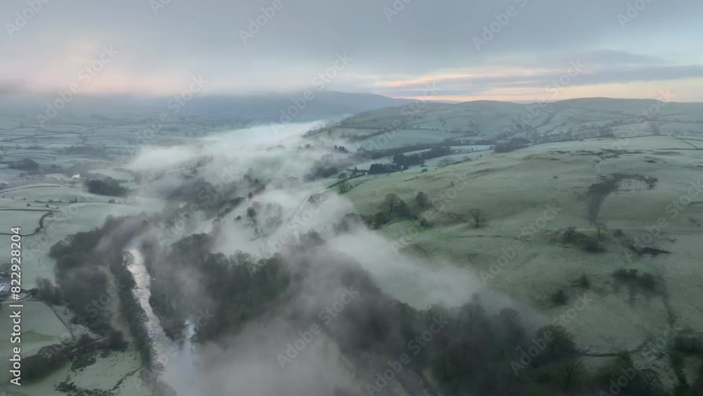Misted patchwork field hillside with river shrouded by mist and dawn glow on the horizon at sunrise in winter. English Lake District, Cumbria, UK. Misted patchwork field hillside with river shrouded by mist and dawn glow on the horizon at sunrise in winter. English Lake District, Cumbria, UK.