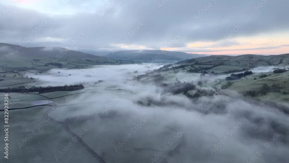 Misty and foggy countryside valley with slow moving cloud static shot at dawn in winter including distant low mountains. Tebay, English Lake District, Cumbria, UK. Misty and foggy countryside valley with slow moving cloud static shot at dawn in winter including distant low mountains. Tebay, English Lake District, Cumbria, UK.