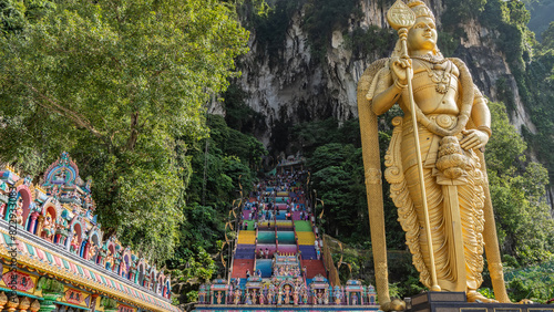 Many tourists climb the rainbow colored stairs to the entrance of the Batu cave. In the foreground is a huge golden statue of the god Lord Murugan, the roof of the temple with colorful sculptures. 