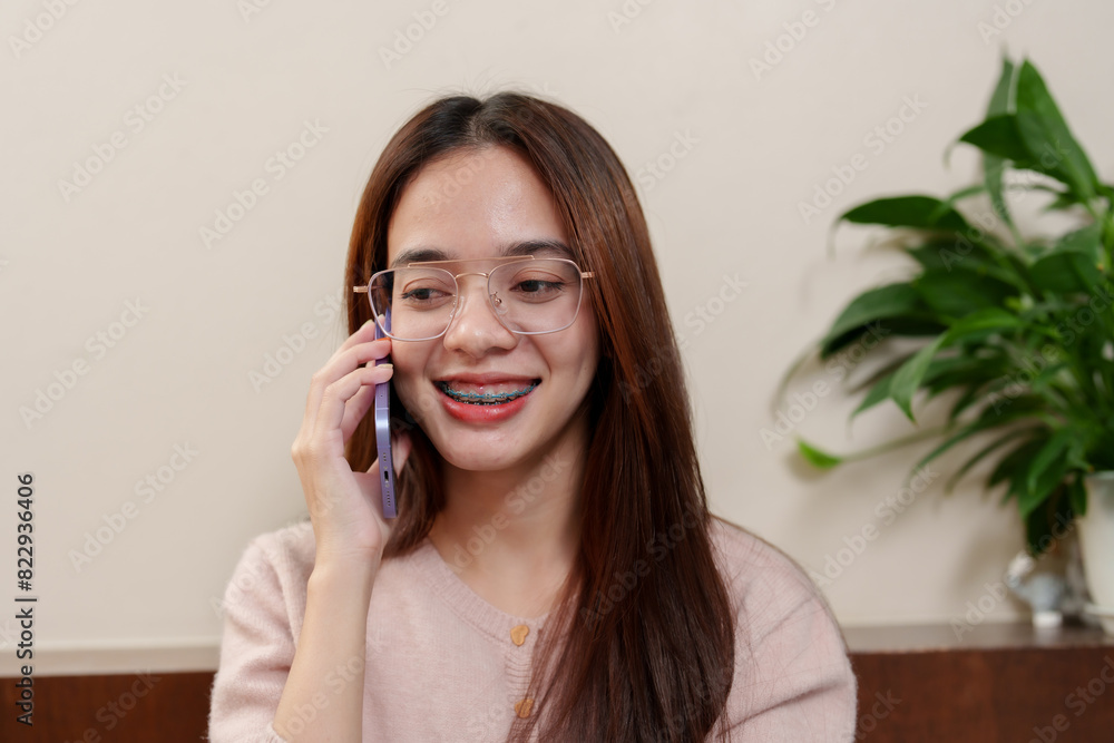 Young Asian girl with braces and glasses, wearing pink sweater, holding smartphone to ear, smiling while talking. Relaxed holiday moment at home with green plants in background. Young Asian girl with braces and glasses, wearing pink sweater, holding smartphone to ear, smiling while talking. Relaxed holiday moment at home with green plants in background.