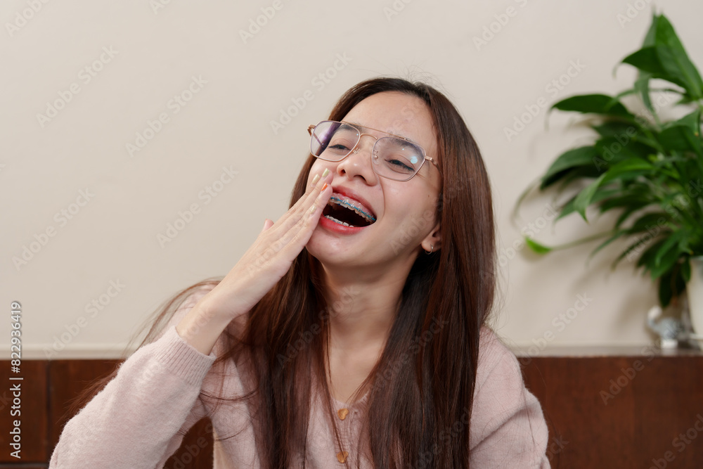 Young Asian girl with braces and glasses, wearing pink sweater, covering mouth while laughing. Relaxed holiday moment at home with green plants in background. Joyful, candid expression. Young Asian girl with braces and glasses, wearing pink sweater, covering mouth while laughing. Relaxed holiday moment at home with green plants in background. Joyful, candid expression.