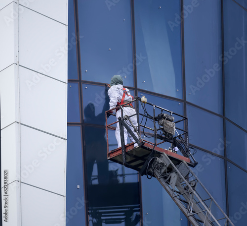 An installer dressed in a uniform and a safety harness cleans the glass facades while standing on a construction winch