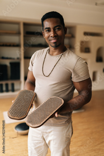 Vertical portrait of positive black male holding in hands wooden Sadhu Board with nails standing in cozy living room, preparing to practice, smiling looking at camera. Concept of mindful meditation