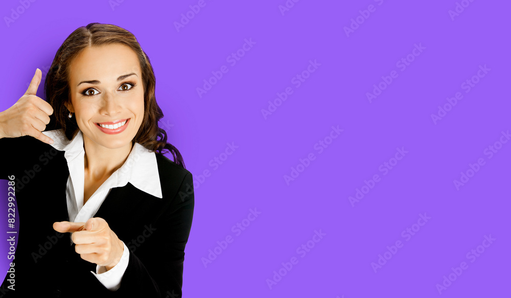 Obraz premium Businesswoman wear black confident suit showing call me hand sign gesture and pointing at viewer, isolated purple wall background. Portrait of smiling brunette woman at studio. Business ad concept.