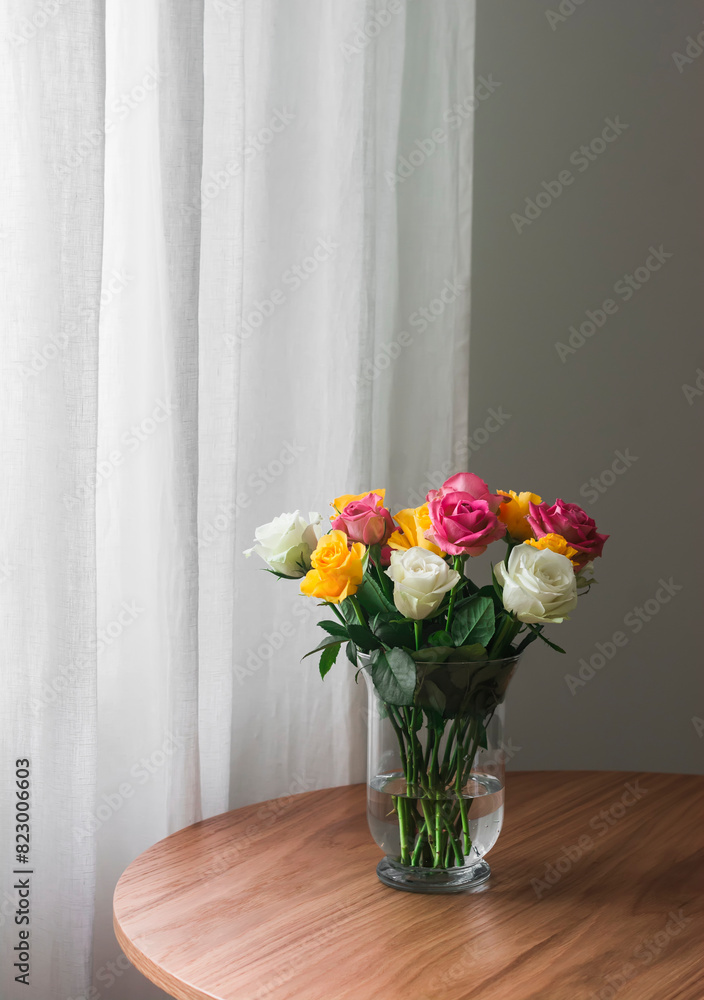 A bright bouquet of yellow, white, pink roses in a glass vase on a wooden table in the living room A bright bouquet of yellow, white, pink roses in a glass vase on a wooden table in the living room
