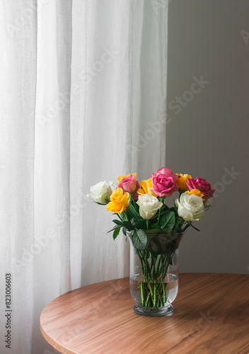 A bright bouquet of yellow  white  pink roses in a glass vase on a wooden table in the living room