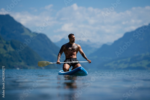 Male fit muscular model with paddle board. Guy paddling on paddleboard. Healthy summer lifestyle. Water sport. SUP surfing in summer vacation in Alps lake in Switzerland.