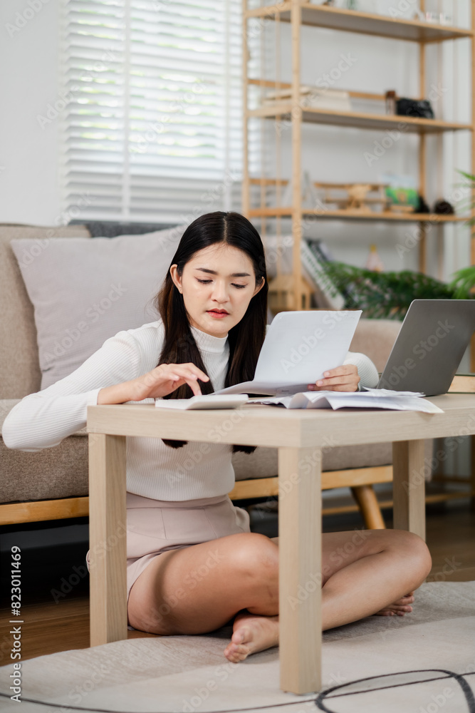 A woman is sitting on the floor in front of a table with a laptop and a stack of papers. She is focused on the papers and seems to be working on something important. The room has a cozy A woman is sitting on the floor in front of a table with a laptop and a stack of papers. She is focused on the papers and seems to be working on something important. The room has a cozy