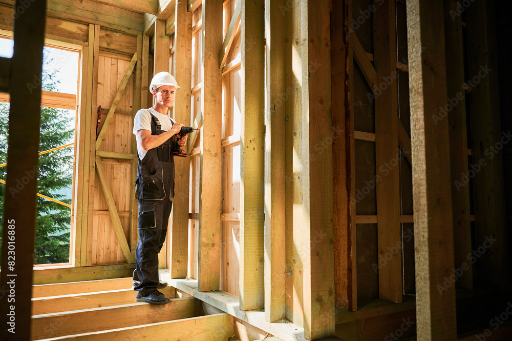 Carpenter constructing wooden framed house. Man worker working with screwdriver, wearing work overalls and helmet. Concept of modern eco-friendly construction. Carpenter constructing wooden framed house. Man worker working with screwdriver, wearing work overalls and helmet. Concept of modern eco-friendly construction.
