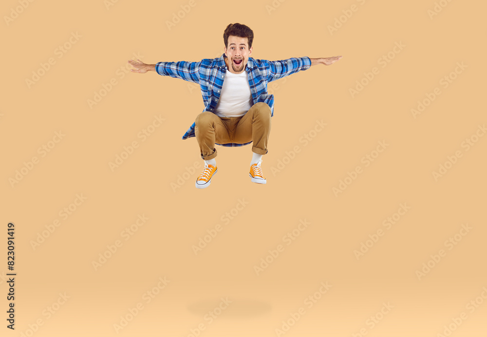Cheerful, active and energetic young man jumping high on beige background in studio. Caucasian guy with bent legs and outstretched arms is levitating in air and laughing at camera. Full length. Cheerful, active and energetic young man jumping high on beige background in studio. Caucasian guy with bent legs and outstretched arms is levitating in air and laughing at camera. Full length.