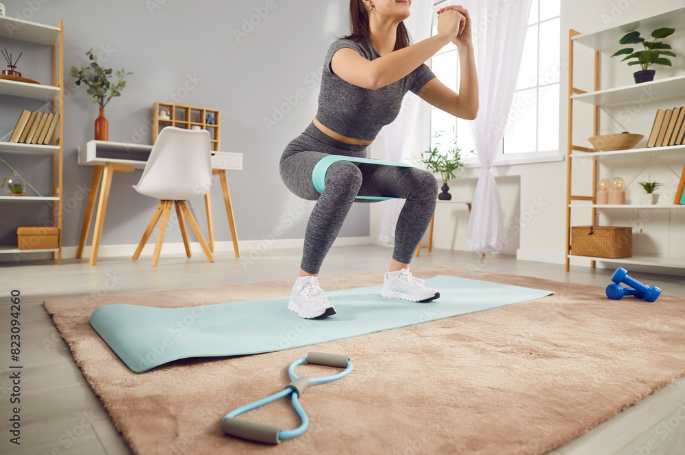 Obraz premium Young sporty woman doing stretching exercises with rubber band standing in living room at home. Happy athletic girl doing domestic workout. Sport, fitness and home training concept.