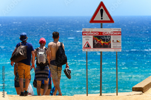 Lagos, Faro, Algarve, Portugal, Europe - group of young people walking down to Pinhao beach, Praia do Pinhao, warning sign - rockfall hazard