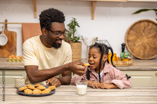 Happy African father and daughter eating cookies and drinking milk kitchen