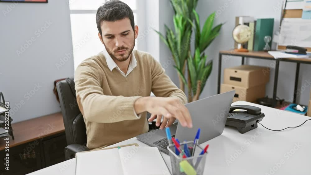 A young hispanic man working in an office, with a laptop, phone, and plant in the background. A young hispanic man working in an office, with a laptop, phone, and plant in the background.