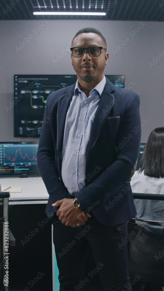 African American IT scientist stands in monitoring room and looks at camera. Multi cultural big data engineers work at computers. Multiple screens with displayed real-time analysis charts. Vertical African American IT scientist stands in monitoring room and looks at camera. Multi cultural big data engineers work at computers. Multiple screens with displayed real-time analysis charts. Vertical