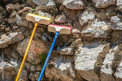 Two colorful cleaning floor brushes with long handle leaning on the stone wall background, out door cleaning concept